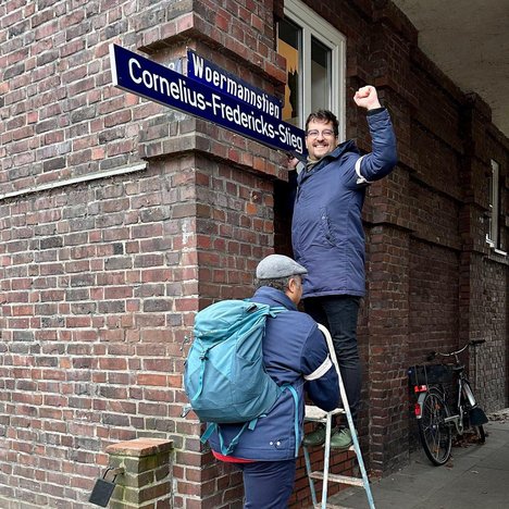 Rachid Messaoudi (Die Linke) auf einer Leiter stehend und ein Straßenschild haltend. Auf diesem steht Cornelius-Fredericks-Stieg.